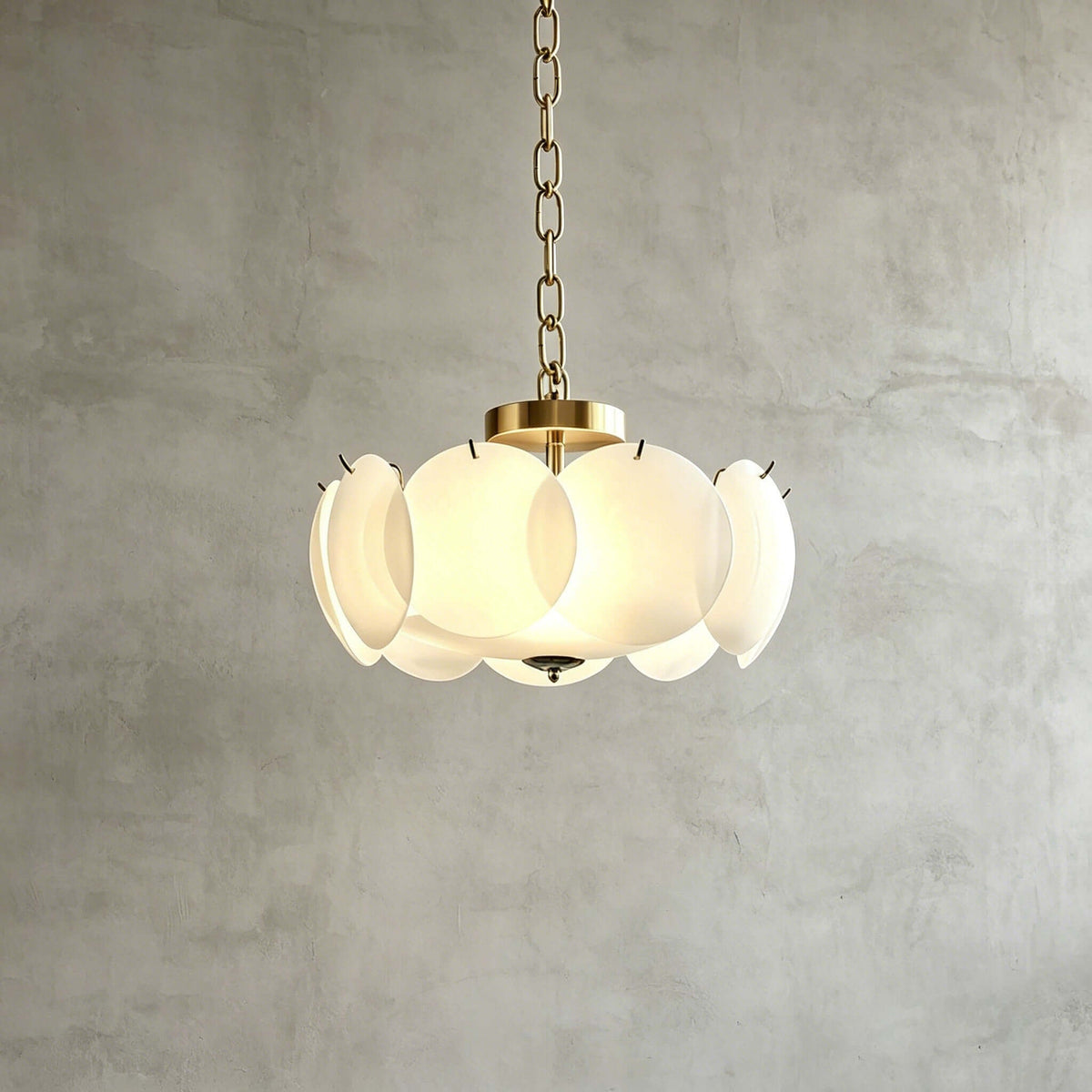 The Clérachic chandelier in brass and white hangs on a brass chain against a grey plaster wall, lit evenly through its glass discs.