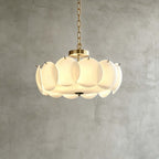 Brass-finish Clérachic chandelier in a wider size, its overlapping white glass discs glowing warmly against a grey plaster wall.
