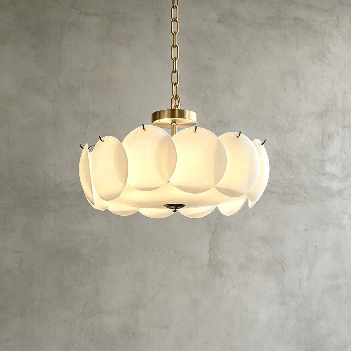 Brass-finish Clérachic chandelier in a wider size, its overlapping white glass discs glowing warmly against a grey plaster wall.