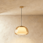 Clérachic larger pendant light in amber glass over a glowing opal inner shade, hung from a brass canopy on a black cord.