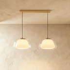 Clérachic two-head pendant light with clear glass shades over opal diffusers, hung from a long brass bar canopy on black cords.