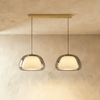 Clérachic two-head pendant light in smoked grey glass with opal diffusers, hung from a brass bar canopy on black cords.