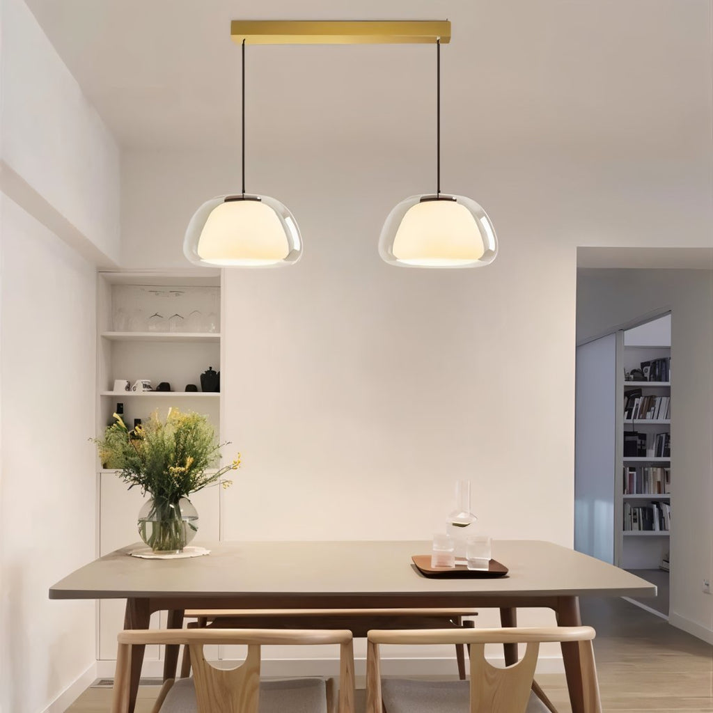 Clérachic two-head pendant light with clear glass shades hung from a brass bar above a wooden dining table with a flower vase.