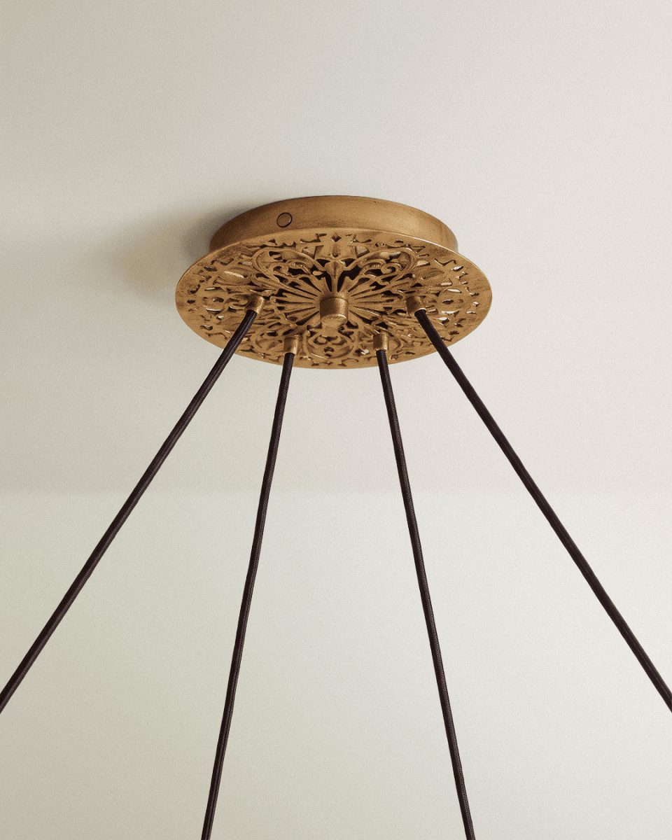 Clérachic chandelier ceiling canopy in aged gold with pierced scrollwork and four dark fabric cords descending from it.