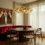 Clérachic gold and glass petal chandelier lit over a round wooden table, red velvet banquette and sheer curtains behind.