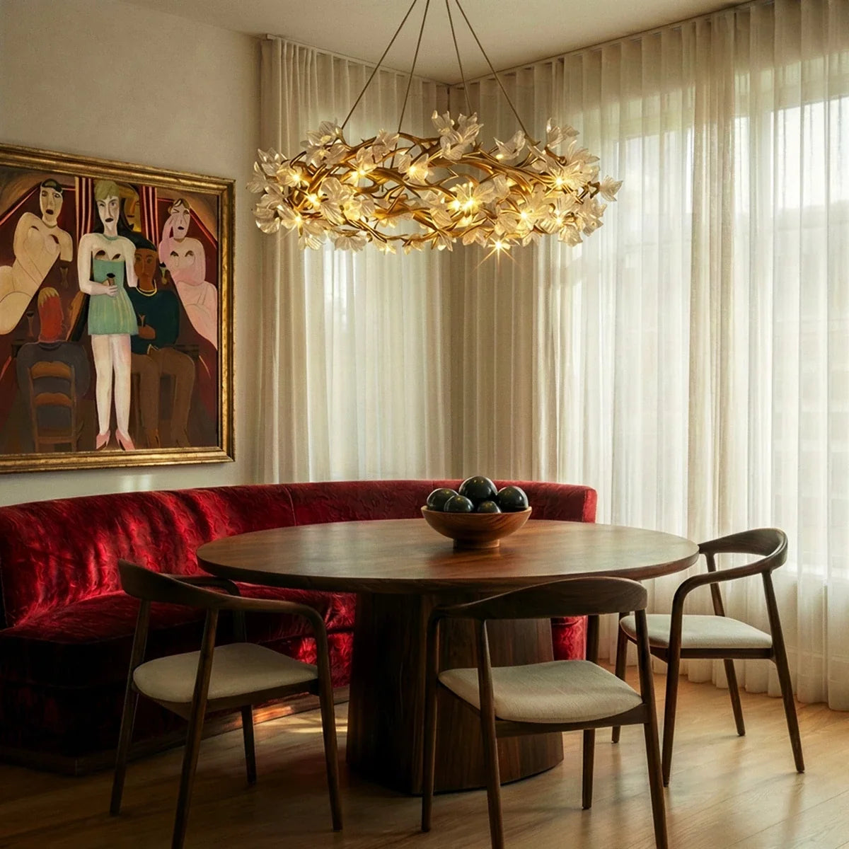 Clérachic gold and glass petal chandelier lit over a round wooden table, red velvet banquette and sheer curtains behind.