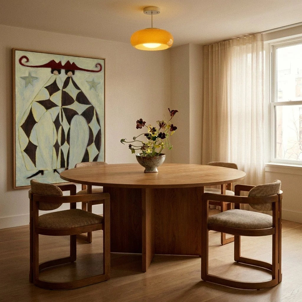 Clérachic yellow glass pendant lit above a round oak dining table, with a bold framed painting and soft curtains behind.