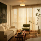 Clérachic white glass pendant glowing above a living room with a cream sofa, green armchair and sheer curtains at golden hour.