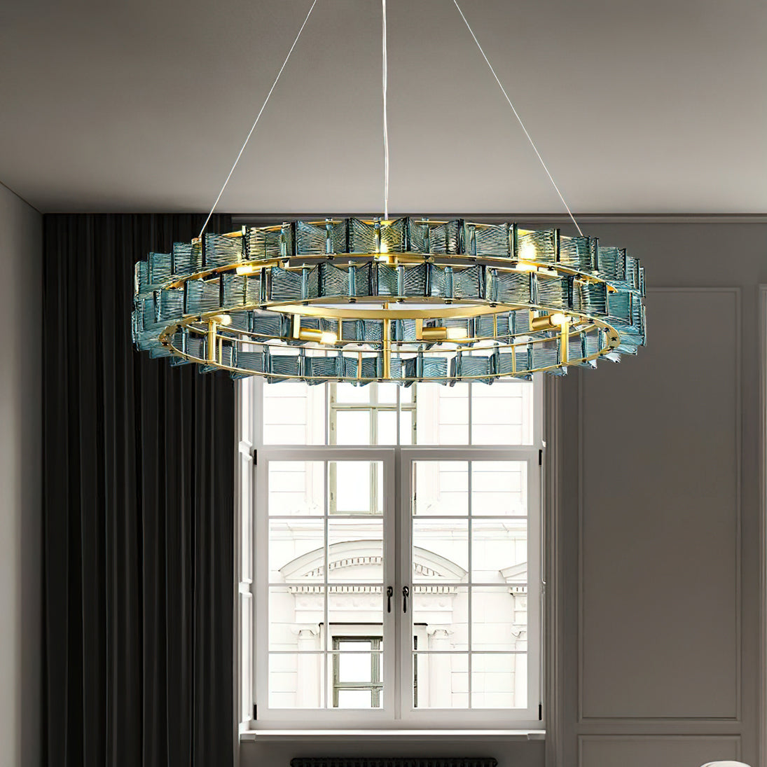 A blue Clérachic chandelier hangs low in front of a white-framed window and dark curtains in a grey room.