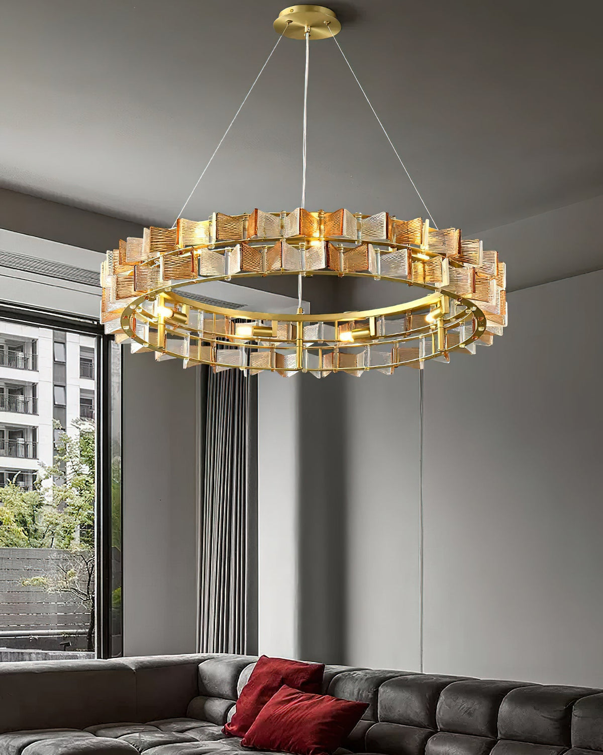 This amber Clérachic chandelier hangs over a dark tufted sofa with red cushions, beside a window onto a city view.
