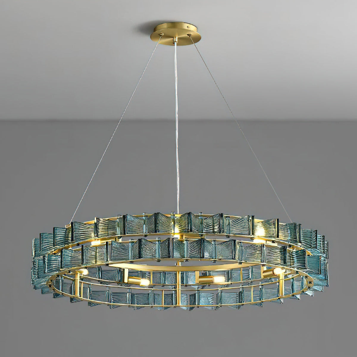 Lit blue Clérachic chandelier on a grey backdrop, its brass ring and spaced bulbs visible beneath two rows of ribbed glass.