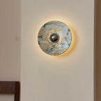 The Clérachic marble wall light lit, with a soft halo of warm light around its edge on a cream wall, its stone gold-veined with dark green.