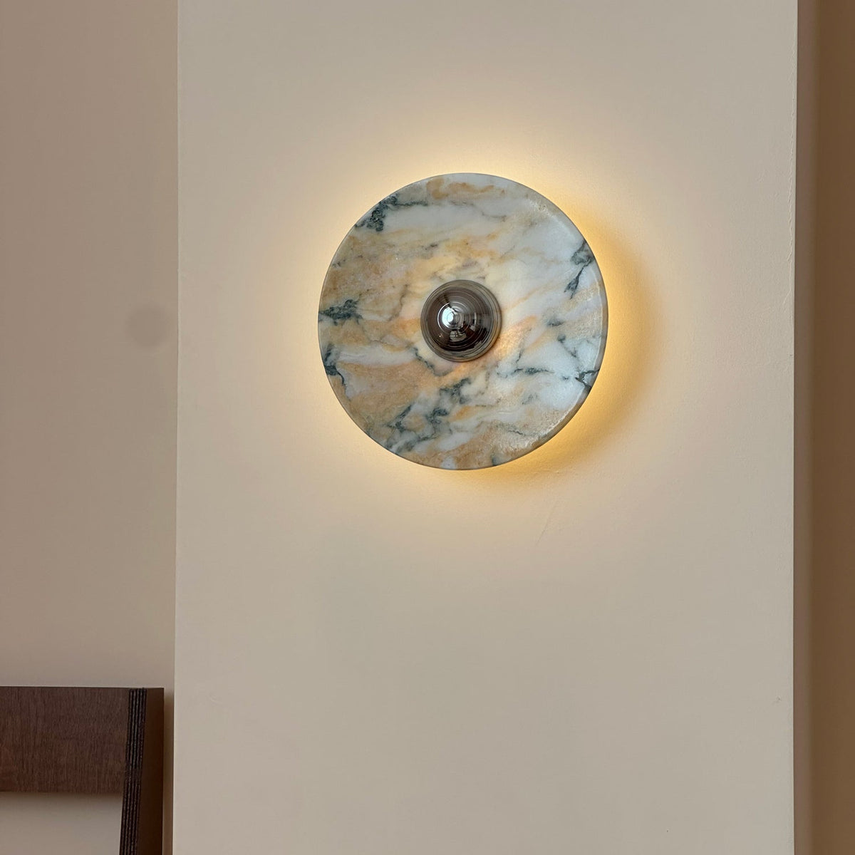 The Clérachic marble wall light lit, with a soft halo of warm light around its edge on a cream wall, its stone gold-veined with dark green.