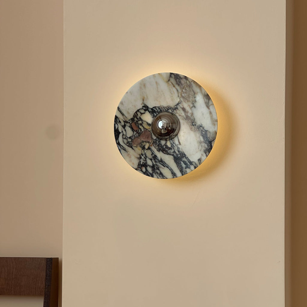A lit Clérachic round marble wall light with dark plum and grey veining and a chrome centre, glowing softly on a cream wall.