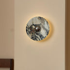 A lit Clérachic round marble wall light with dark plum and grey veining and a chrome centre, glowing softly on a cream wall.