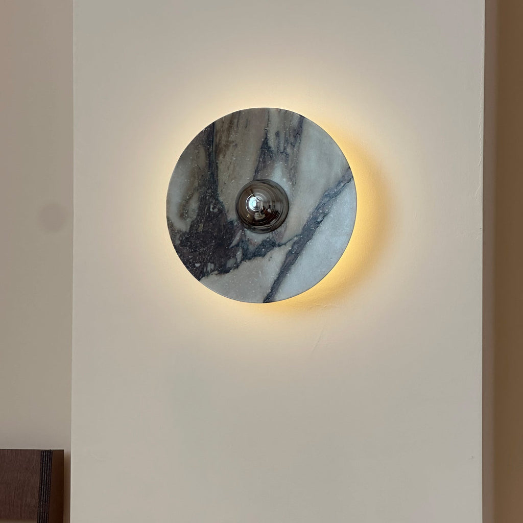 A Clérachic marble wall light in blue-grey stone with dark streaks, with warm light spilling behind its edge onto a pale wall.