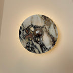 A larger Clérachic marble wall light lit on a cream wall, with a chrome dome at the centre and bold black-and-burgundy veining.