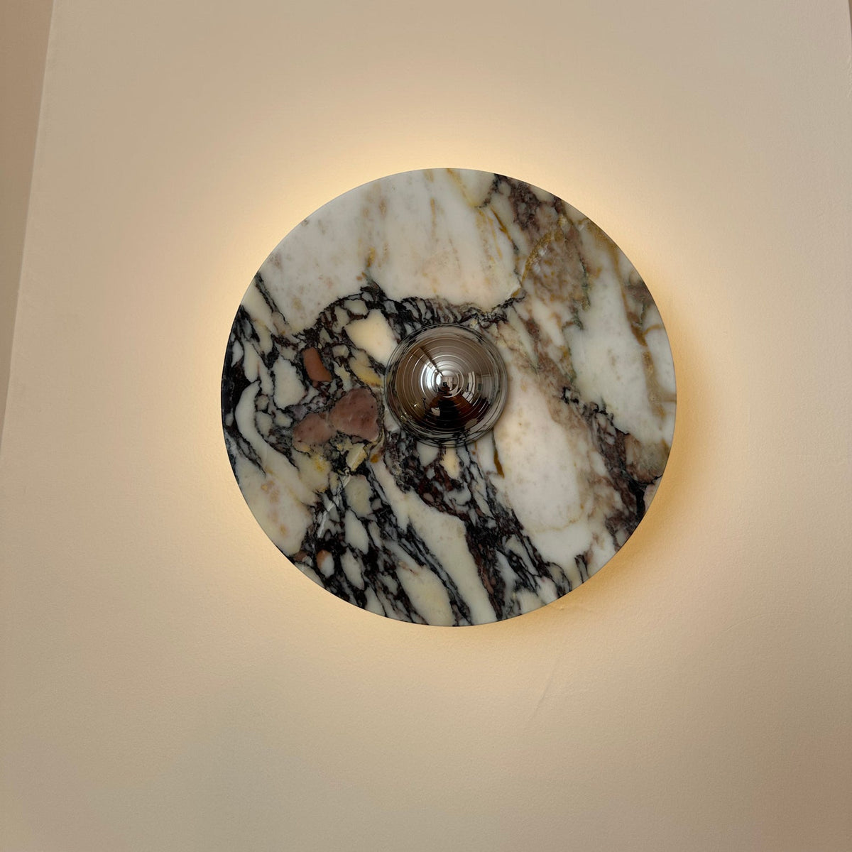 A larger Clérachic marble wall light lit on a cream wall, with a chrome dome at the centre and bold black-and-burgundy veining.