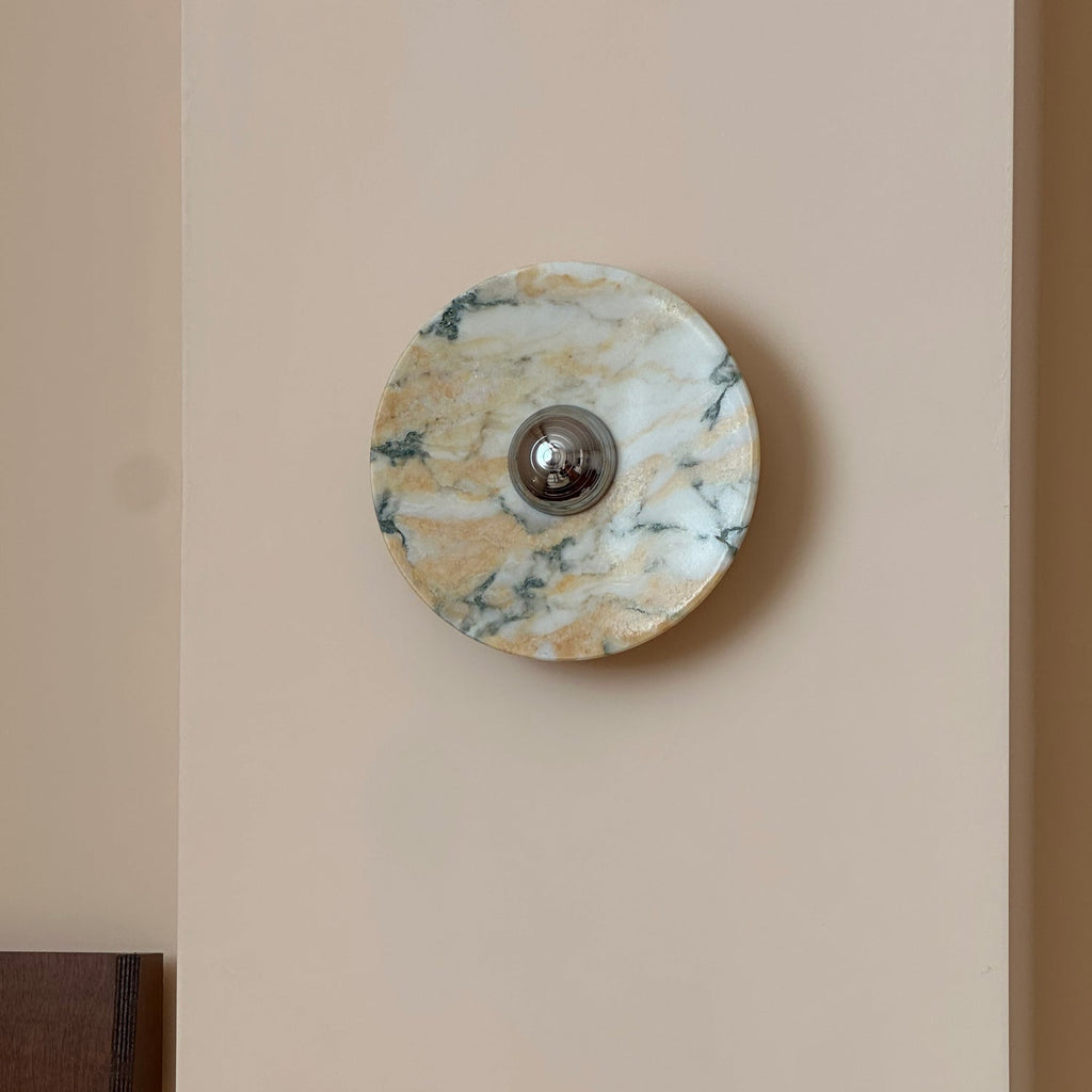 A Clérachic marble wall light switched off, its stone warm gold and white with fine green veins, on a peach-cream wall.