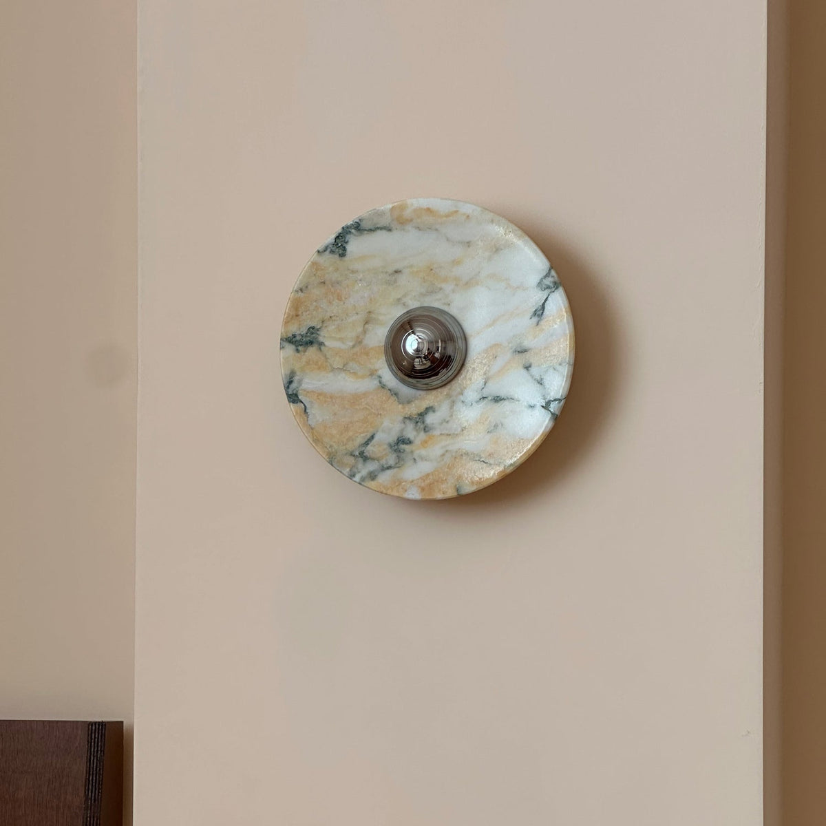 A Clérachic marble wall light switched off, its stone warm gold and white with fine green veins, on a peach-cream wall.
