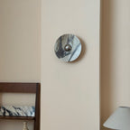 A small Clérachic marble wall light in grey and white stone with dark streaks, high on a cream wall above a lamp shade and a chair back.