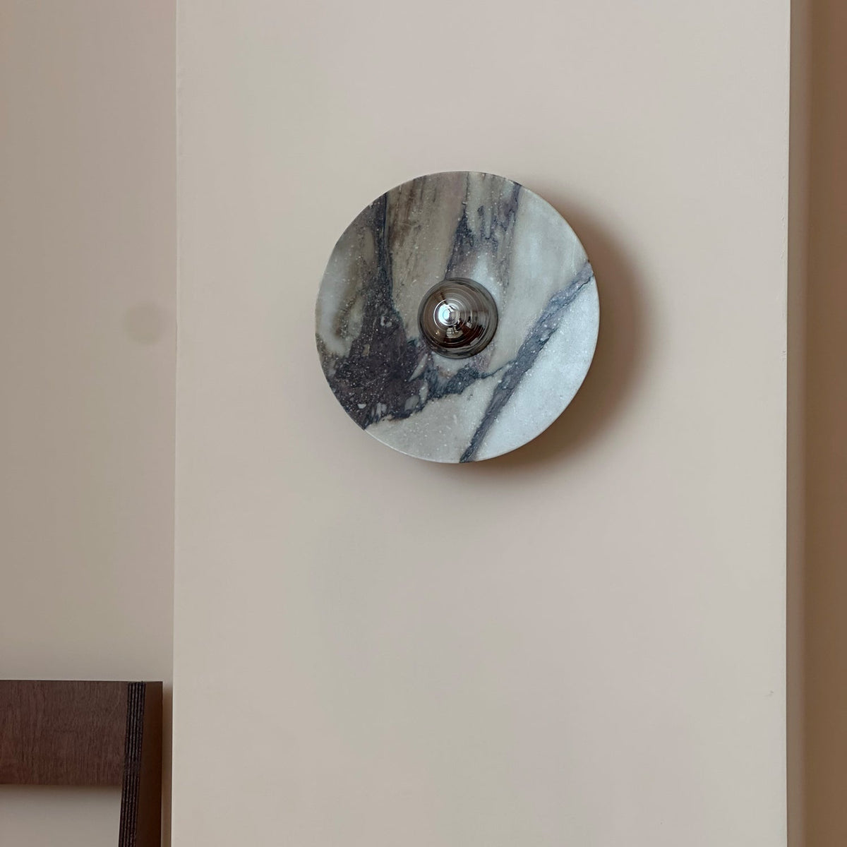 A Clérachic wall light in pale grey marble with black and plum veins, with a chrome dome at the centre, unlit on a pale wall.