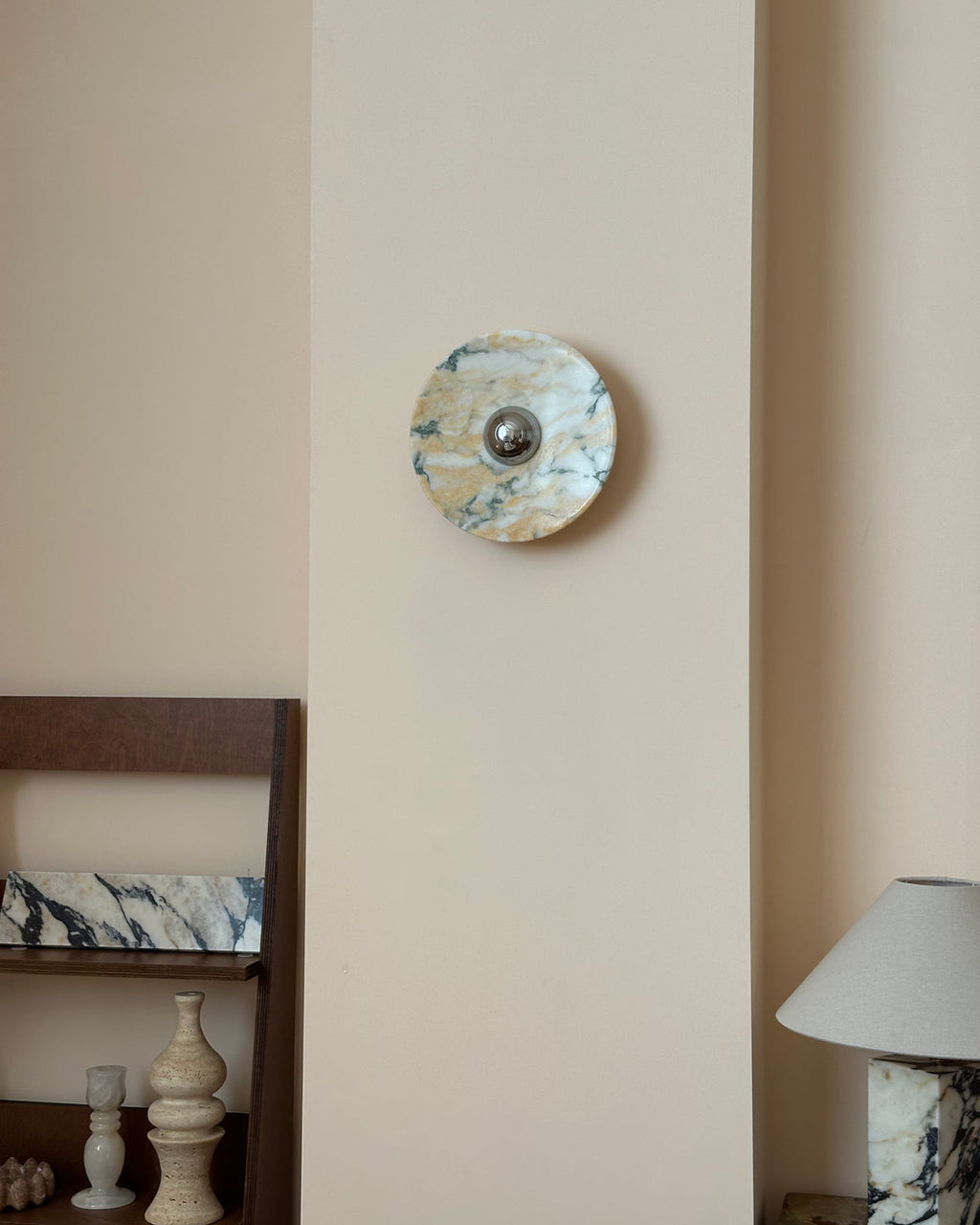 A small Clérachic marble wall light on a tall cream wall panel, above a wooden shelf with stone vases and a marble-base lamp.