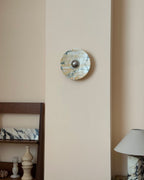 A small Clérachic marble wall light on a tall cream wall panel, above a wooden shelf with stone vases and a marble-base lamp.
