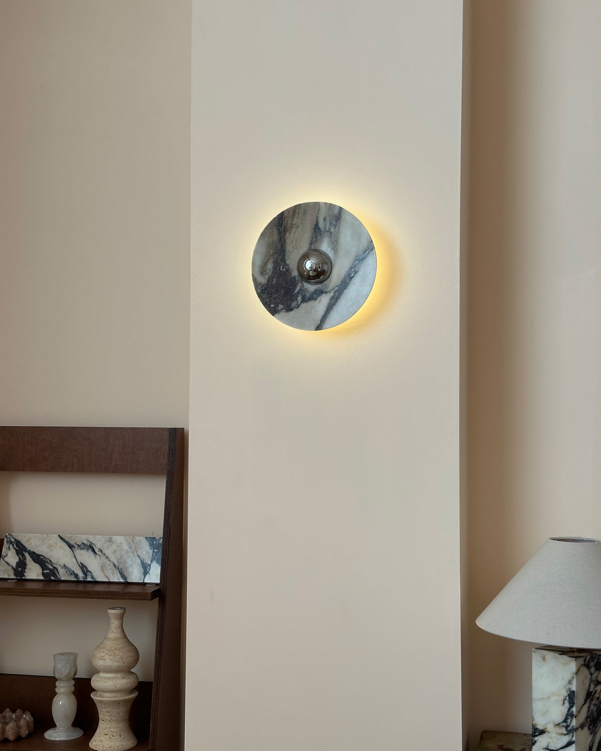 A Clérachic marble wall light lit, with a thin gold halo behind its edge, beside a wooden ladder shelf and a stone-based lamp.