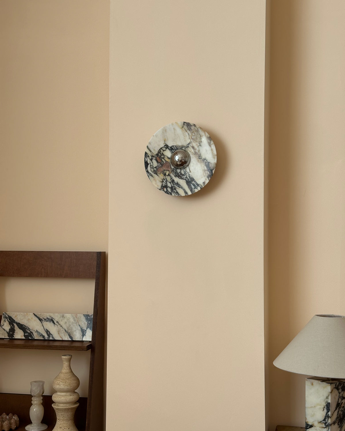 A Clérachic marble wall light unlit on a cream panel, with dark veins and cream patches around its chrome dome and shelving below.