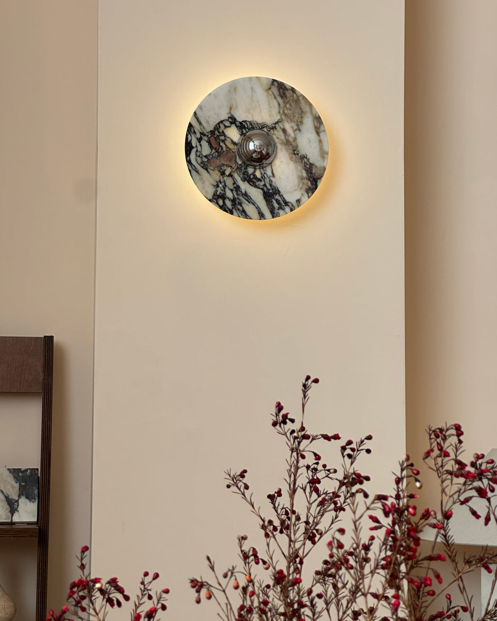 A lit Clérachic marble wall light above a bare branch with red berries, with a glow around the stone's edge on a cream wall.