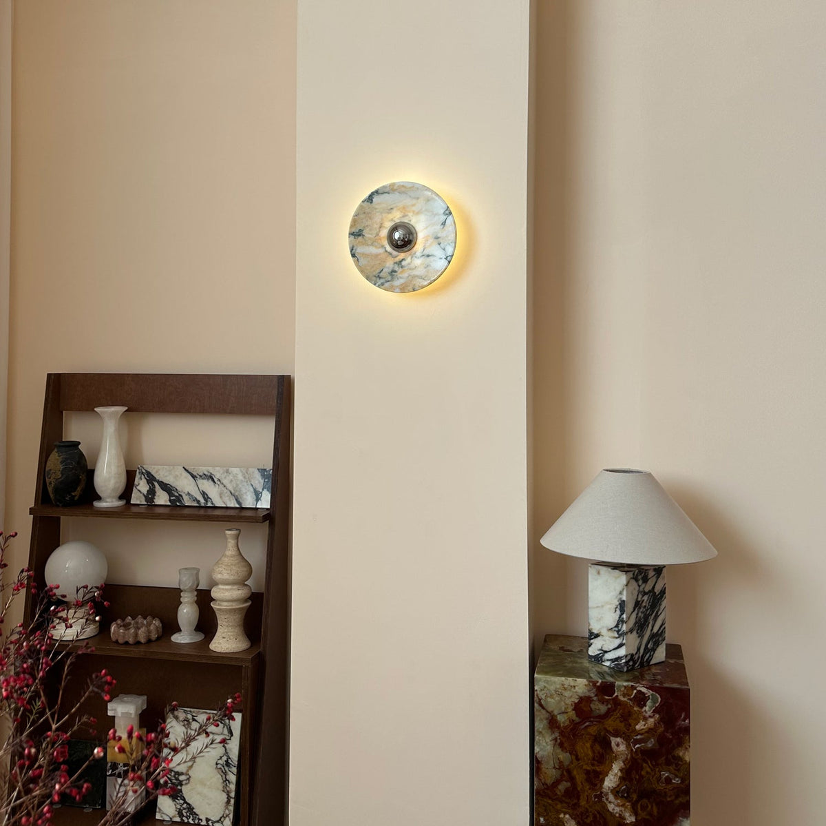A lit Clérachic marble wall light on a cream panel, with a ladder shelf of stone vases and a marble lamp on a green onyx block.