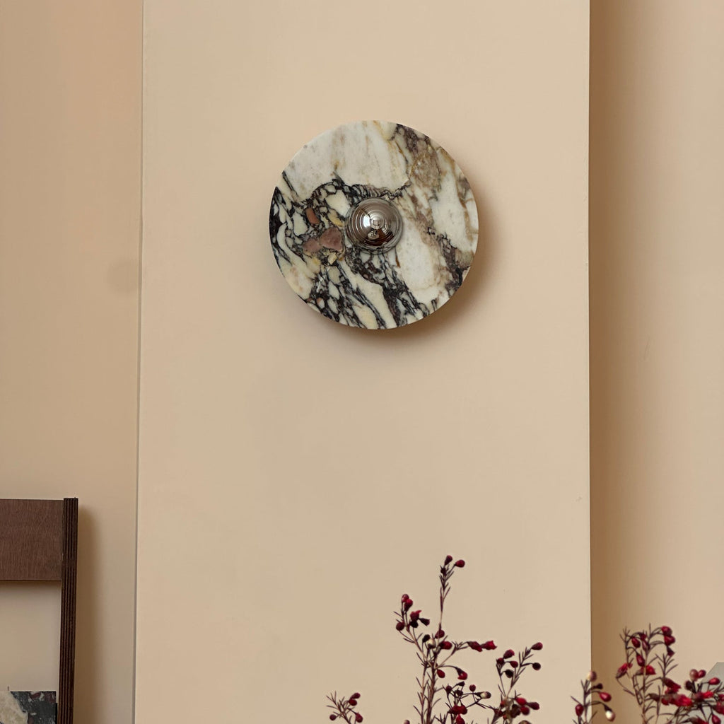 A Clérachic marble wall light unlit on a pale wall, with dark veins on cream stone and red berry branches in the corner.