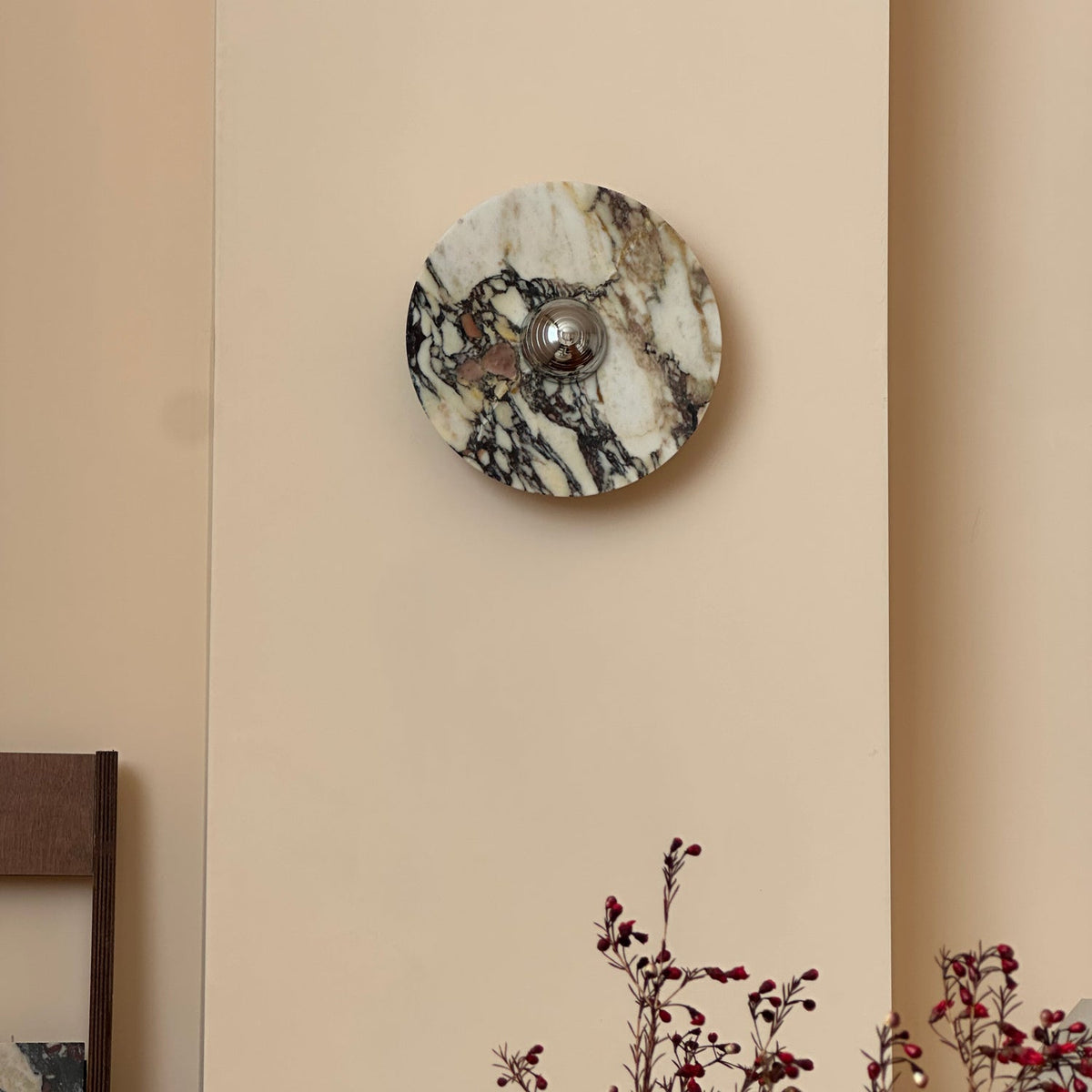 A Clérachic marble wall light unlit on a pale wall, with dark veins on cream stone and red berry branches in the corner.