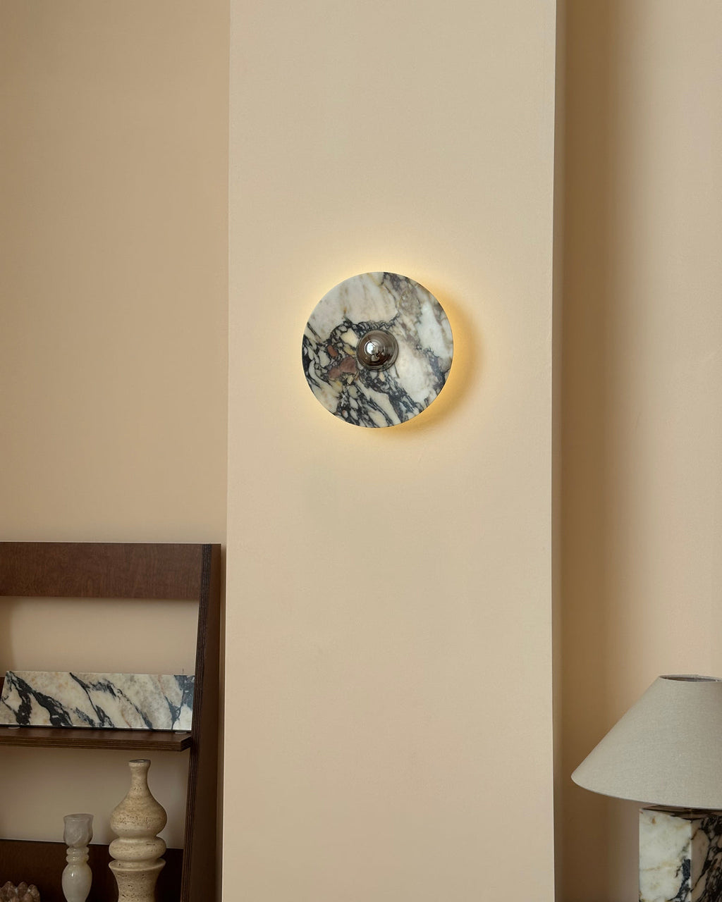 A small Clérachic marble wall light lit with a gentle halo, set above a shelf of vases and a stone-base lamp.