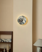 A small Clérachic marble wall light lit with a gentle halo, set above a shelf of vases and a stone-base lamp.