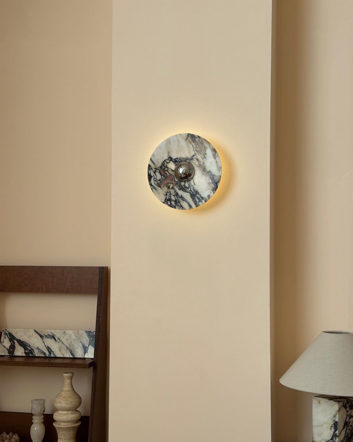 A small Clérachic marble wall light lit with a gentle halo, set above a shelf of vases and a stone-base lamp.