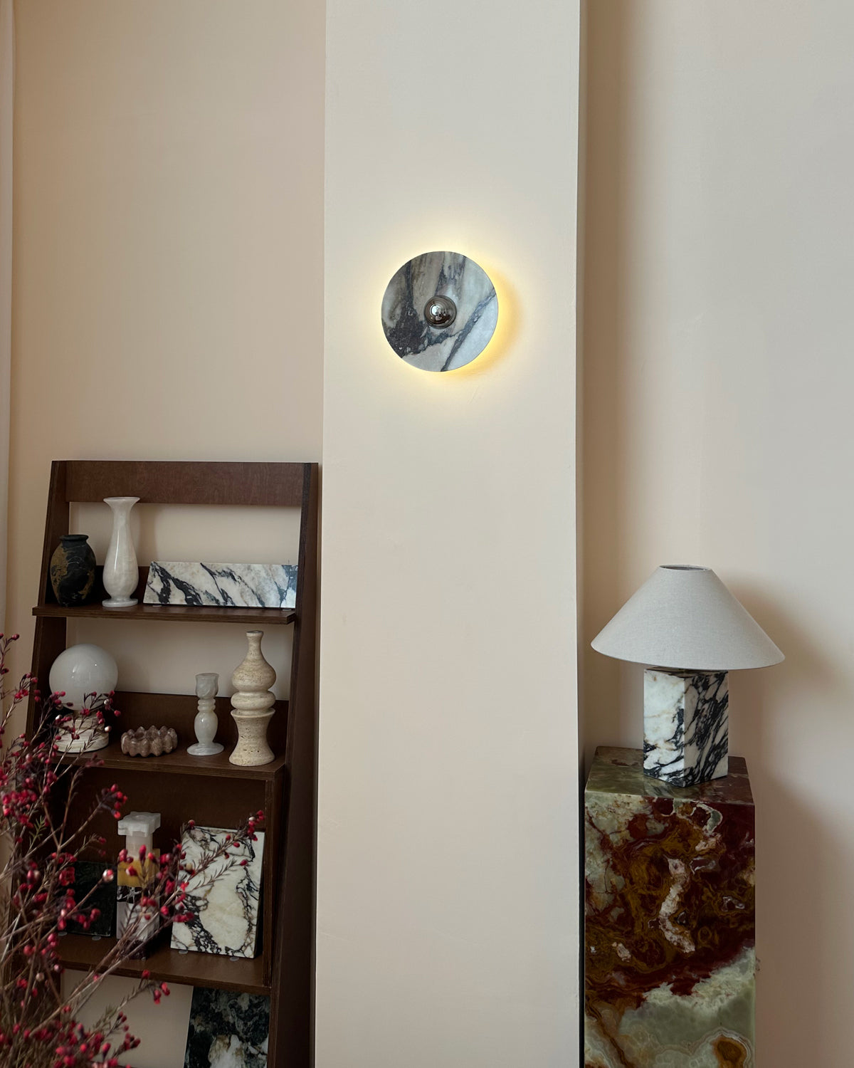 A lit Clérachic marble wall light on a tall cream wall panel, above a ladder shelf of stone pieces and a lamp on a block of green onyx.