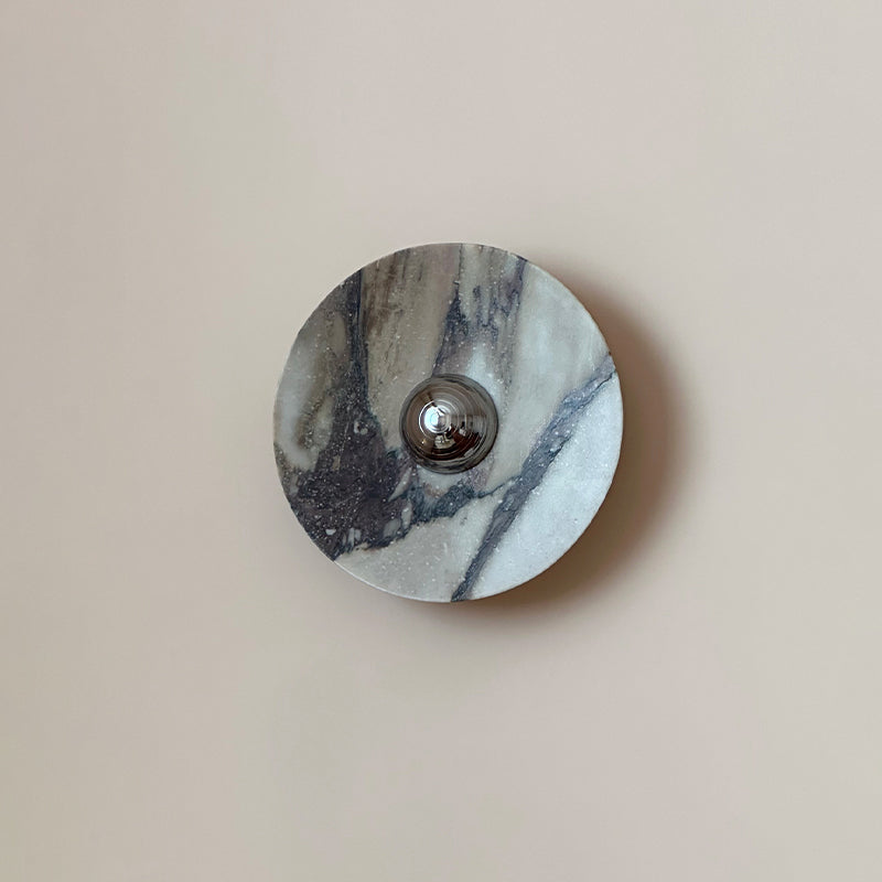 A Clérachic marble wall light in pale grey stone, with one bold diagonal vein across it and a chrome dome centred on a plain pink-beige wall.