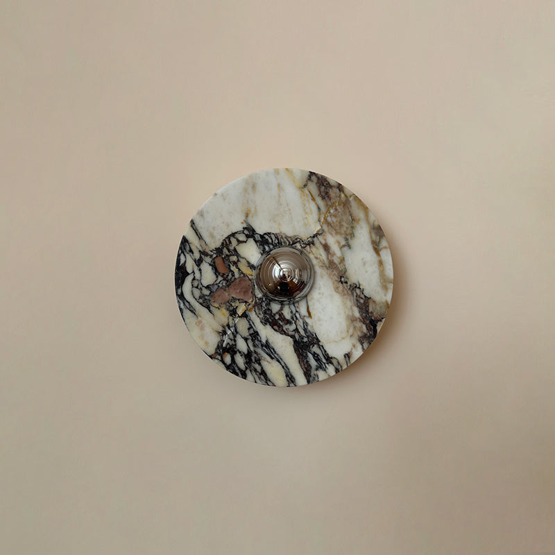 A Clérachic marble wall light in cream stone with black and plum veining and a chrome centre, shown flat on a plain pink-beige wall.