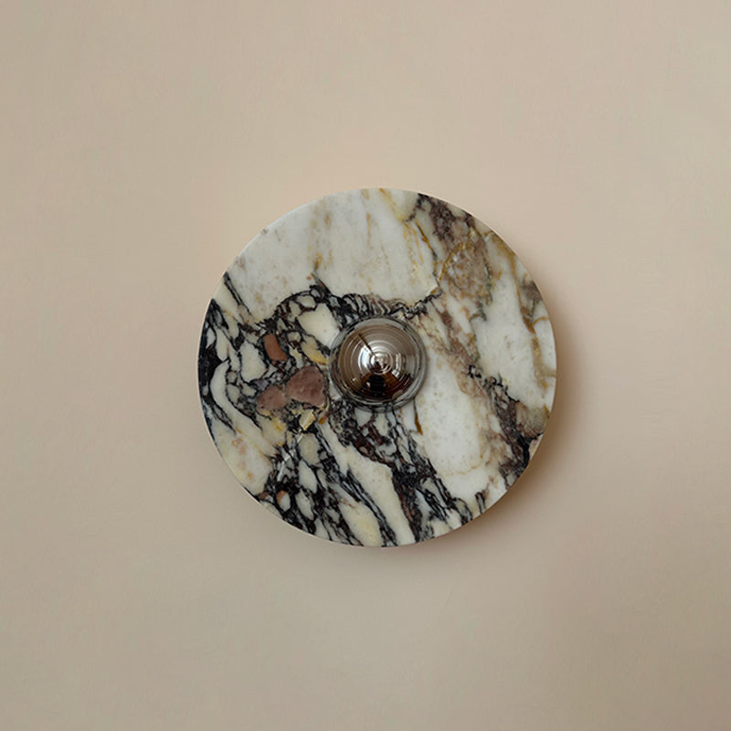 A Clérachic marble wall light with plum-brown patches and black veining across cream stone, on a plain pink-beige wall.