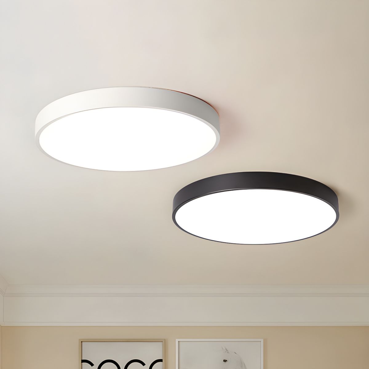 A white and a black round Clérachic ceiling light side by side on a cream ceiling above two framed prints.