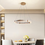 A slim Clérachic travertine ring pendant hangs over a round dining table with fruit and white flowers, in front of a lit bookshelf.