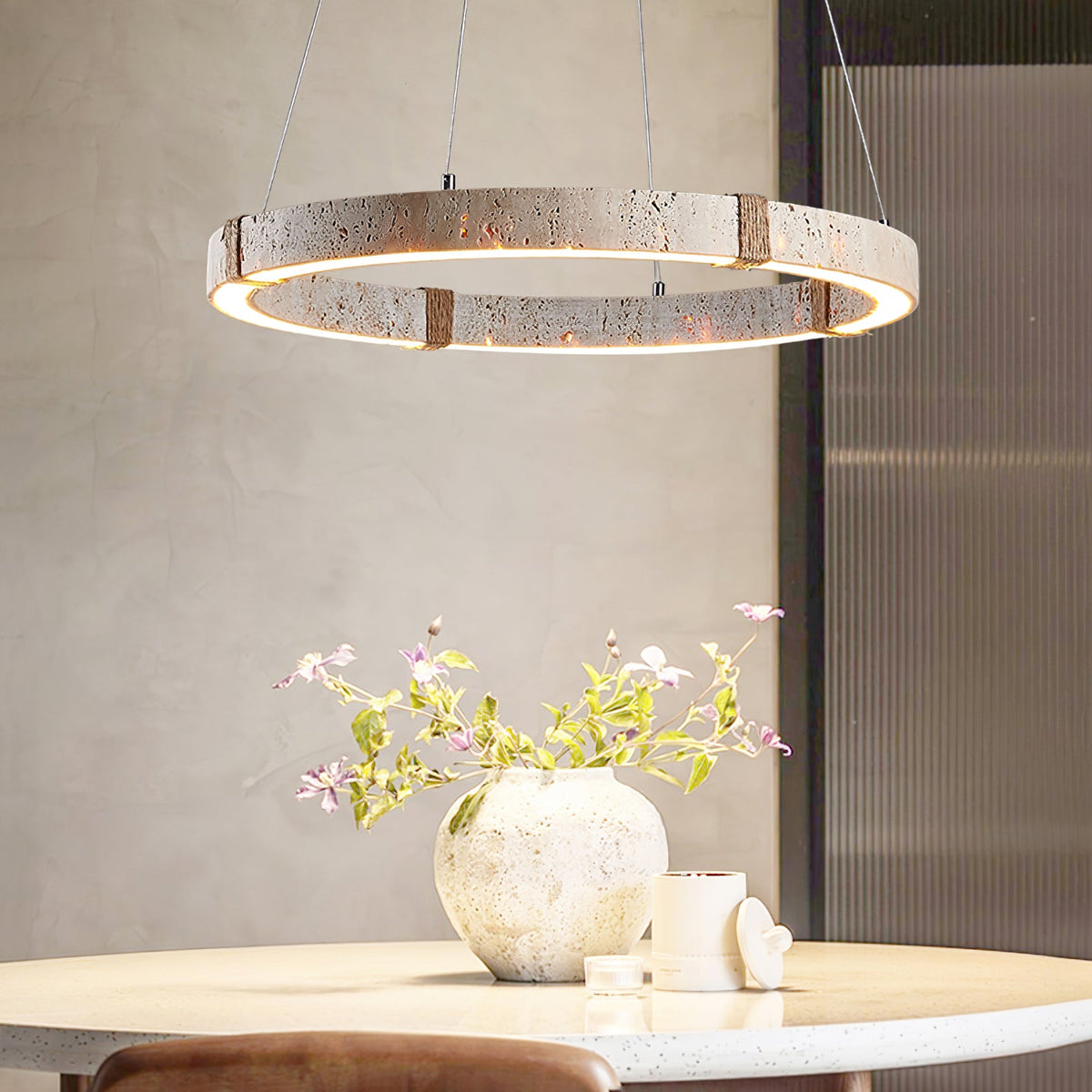 Close view of the travertine ring and warm LED edge of this Clérachic pendant, above a table with a textured vase and pink blossom.