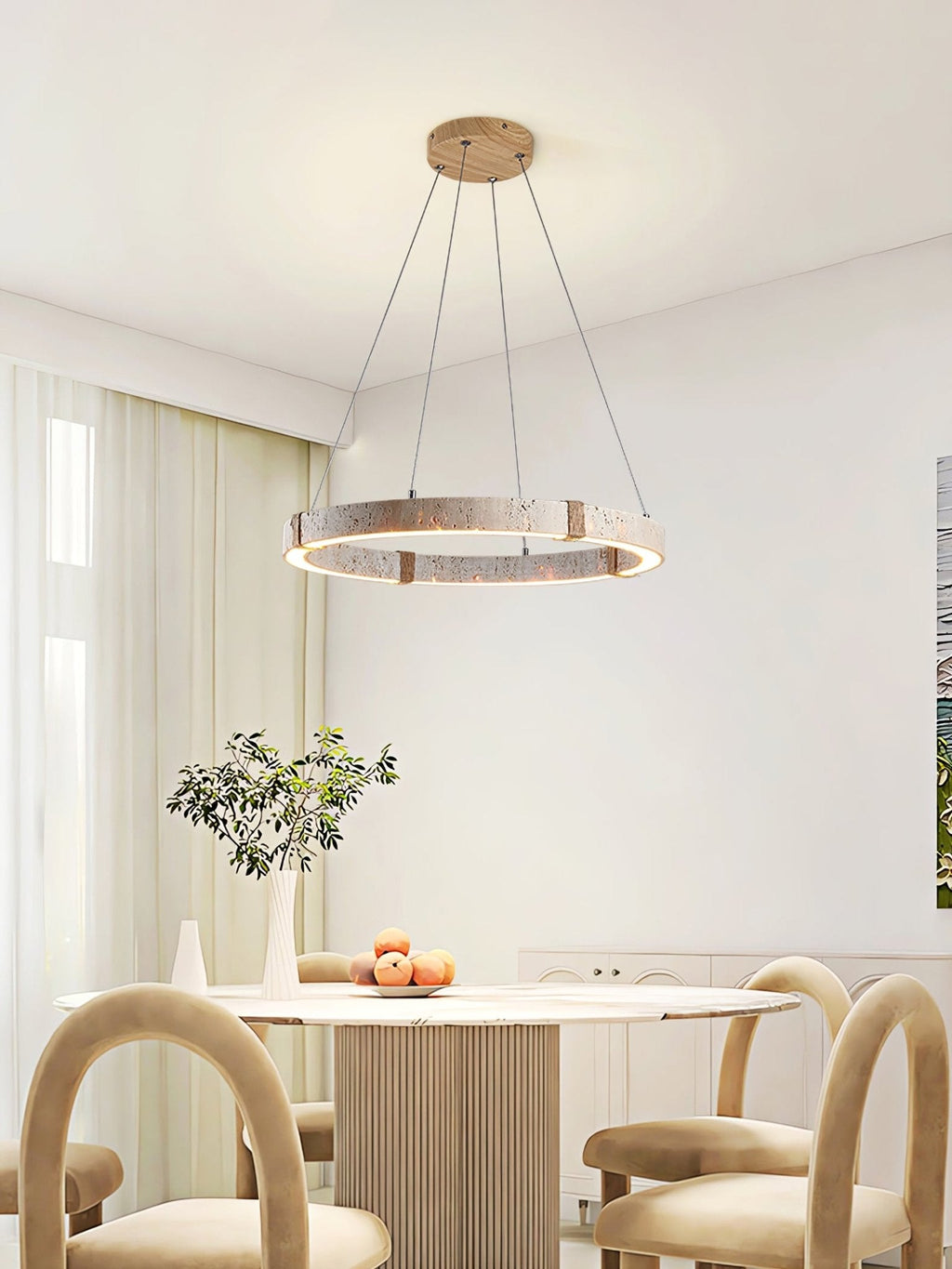 Travertine ring pendant from Clérachic, lit, hanging in a pale dining room above a round table with tan chairs and an olive branch.