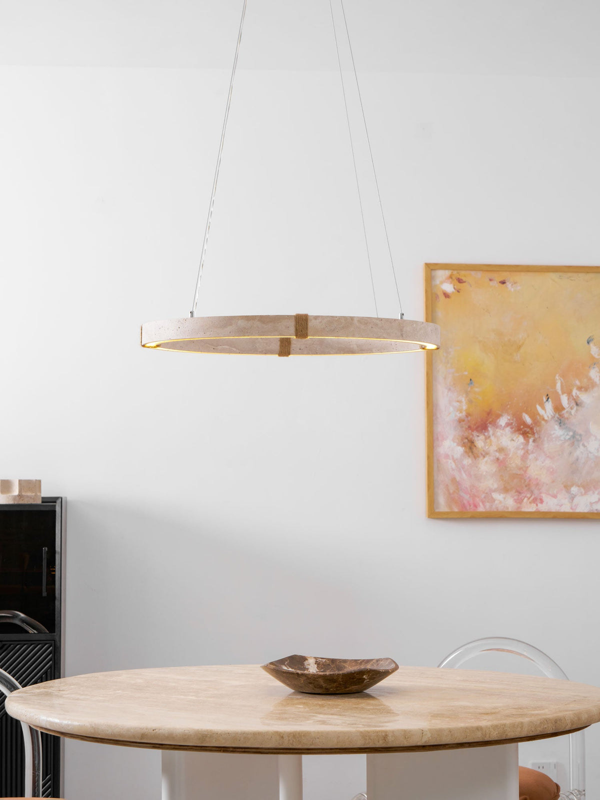 A slim travertine ring pendant from Clérachic hangs above a round travertine table with a dark bowl, beside a large abstract painting.