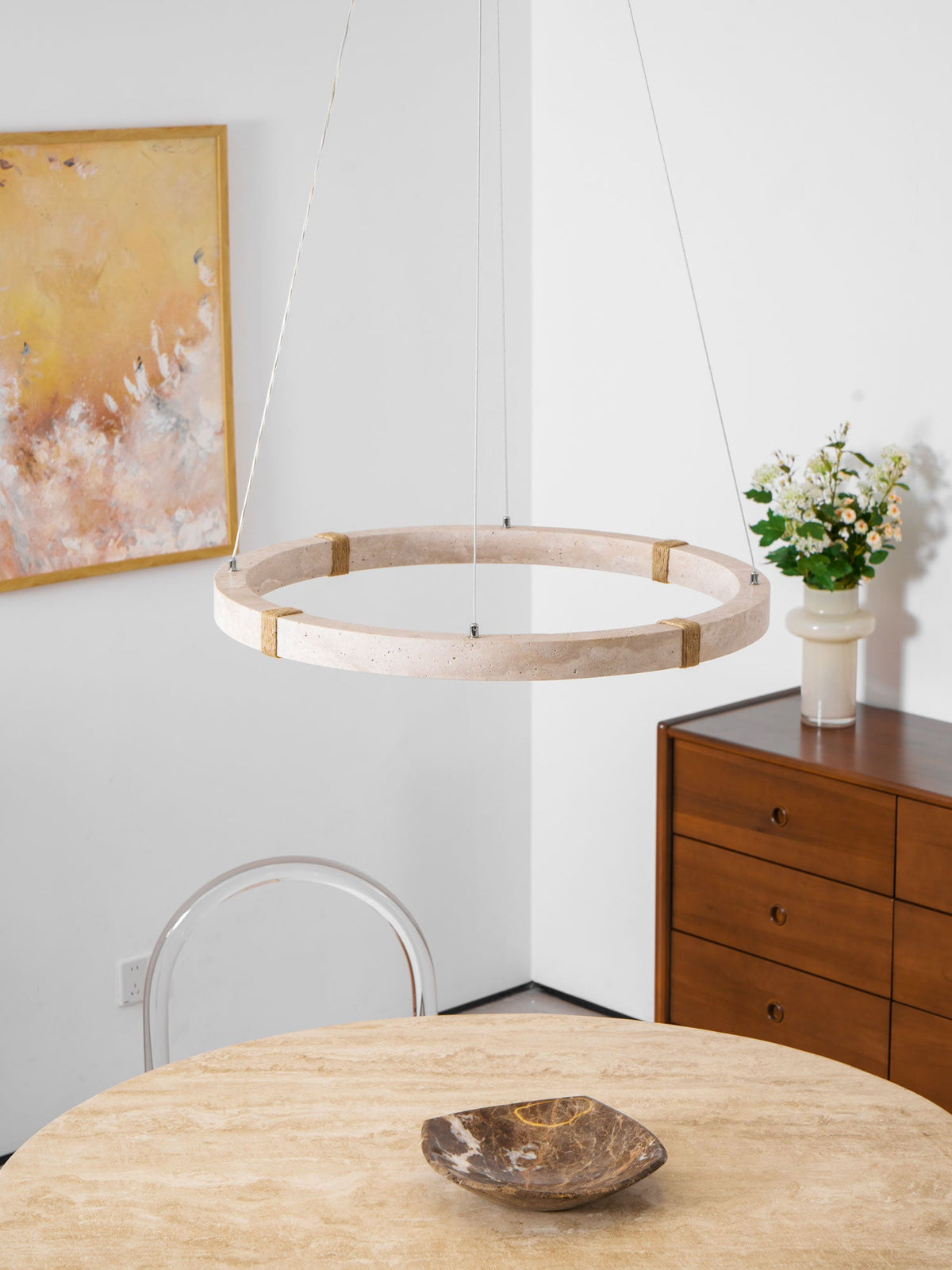 Clérachic travertine ring pendant hung in front of an abstract painting, above a stone-topped table with a dark bowl and a chest of drawers.
