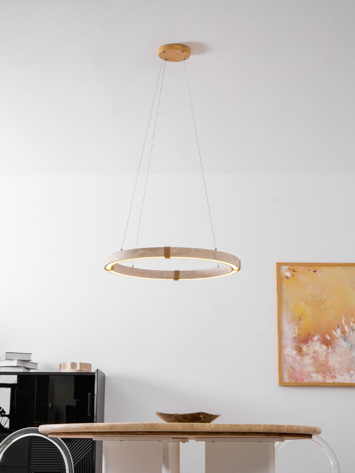 Small travertine ring pendant from Clérachic, lit, in a white room above a round travertine table, beside a painting in warm yellows and pinks.