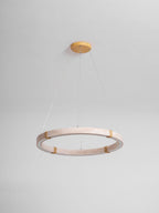 Travertine ring pendant from Clérachic in a pale pink-beige stone, unlit, hung from a gold ceiling plate against a pale grey wall.