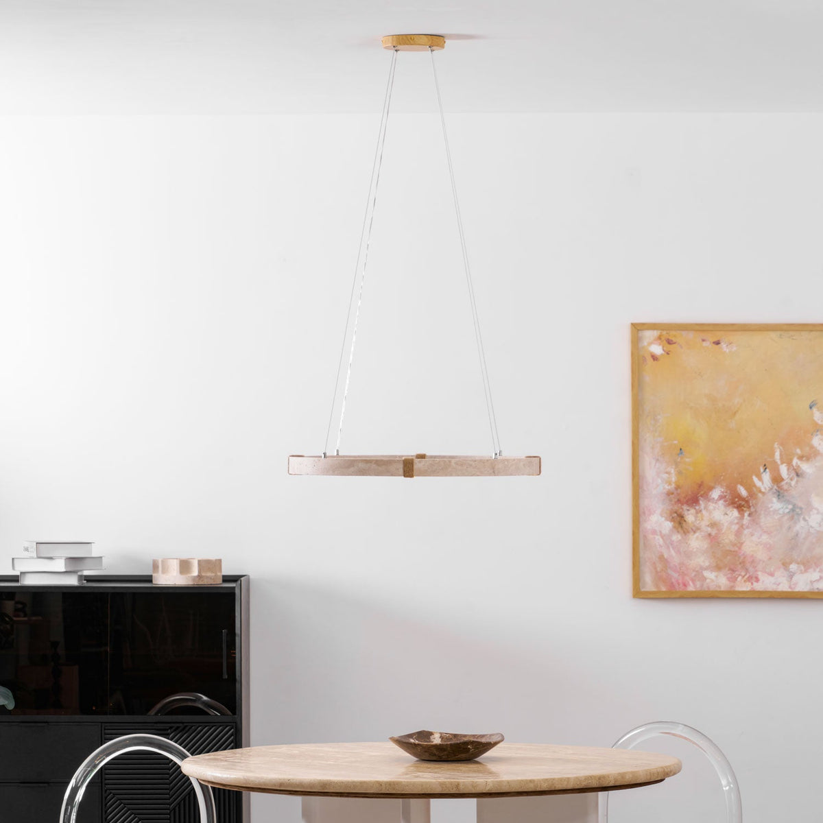 From a distance, a slim Clérachic travertine ring pendant hangs in a white room with a painting and a black cabinet.